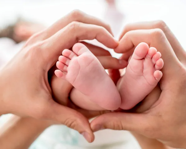 Two hands positioned in a heart shape around a baby's feet