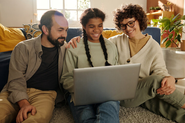A child with braids smiling at a laptop surrounded by her supportive parents