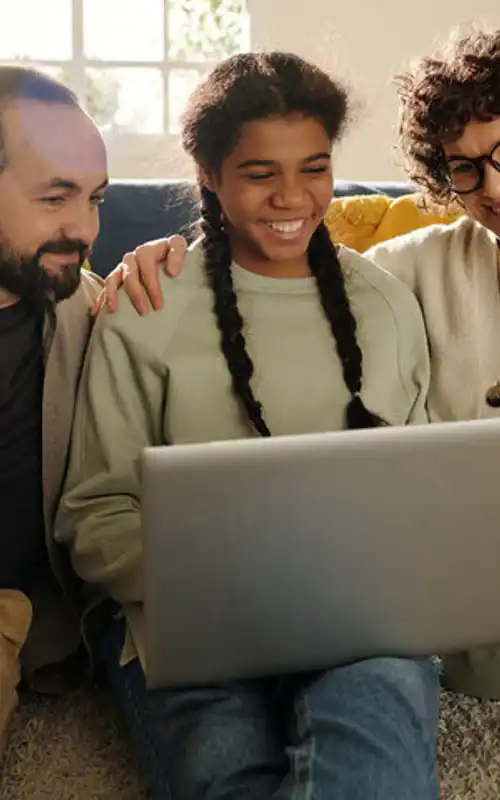 A child with braids smiling at a laptop surrounded by her supportive parents