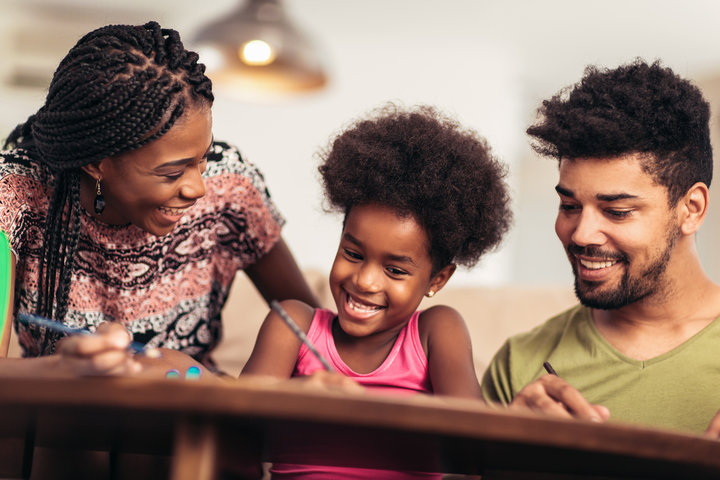An African American family smiling at their child who is holding a pencil and smiling.